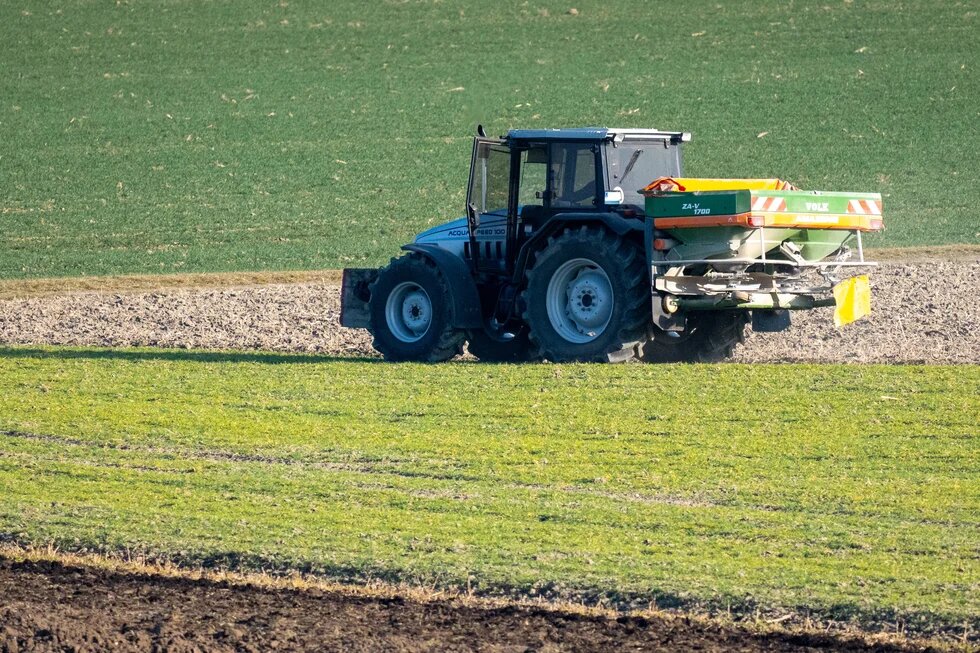 Tractor with rear spreader drives across field, dispersing material; layered farmland stretches into background.