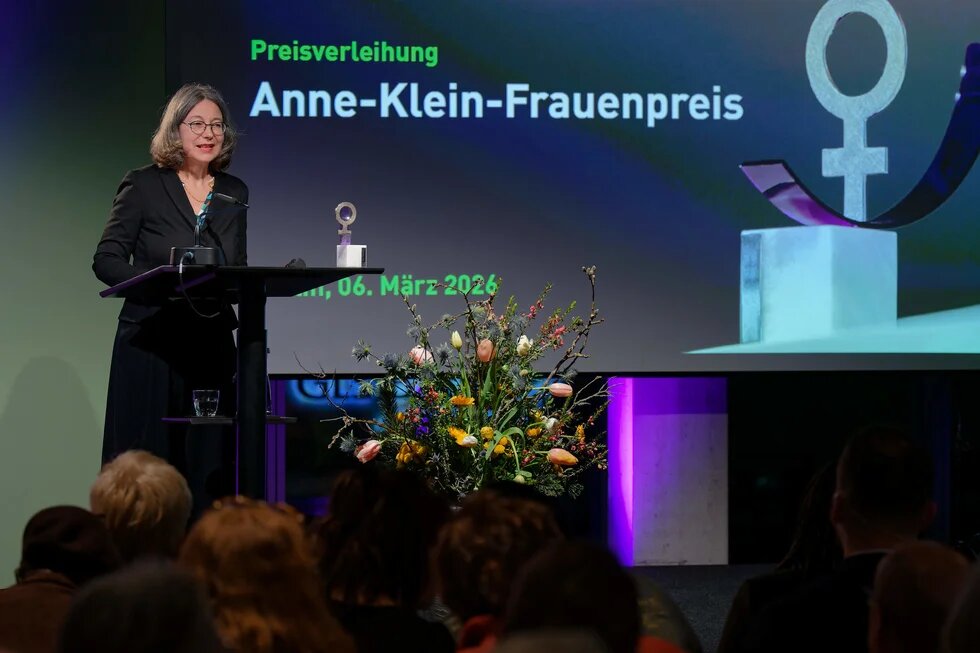 Imme Scholz speaks at the podium in front of an audience; on the screen: the Anne Klein Women’s Award ceremony. A bouquet of flowers and a trophy are visible.