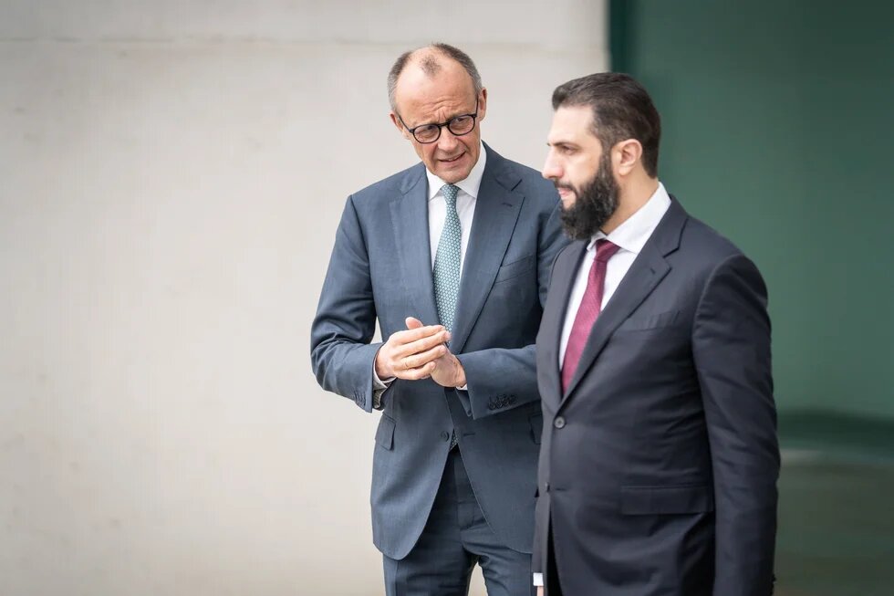 Zwei Männer* in Anzügen gehen nebeneinander und sprechen; einer gestikuliert mit den Händen vor einer Wand.