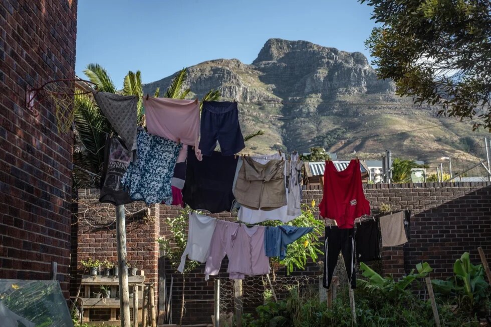 Laundry hangs on lines in a courtyard with brick walls and plants; a large mountain rises in the background under a clear sky.