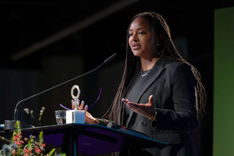 Aminata Touré - A person with long braids is speaking at the podium, gesturing with their hands; a microphone, a glass of water, and a trophy are on the table.