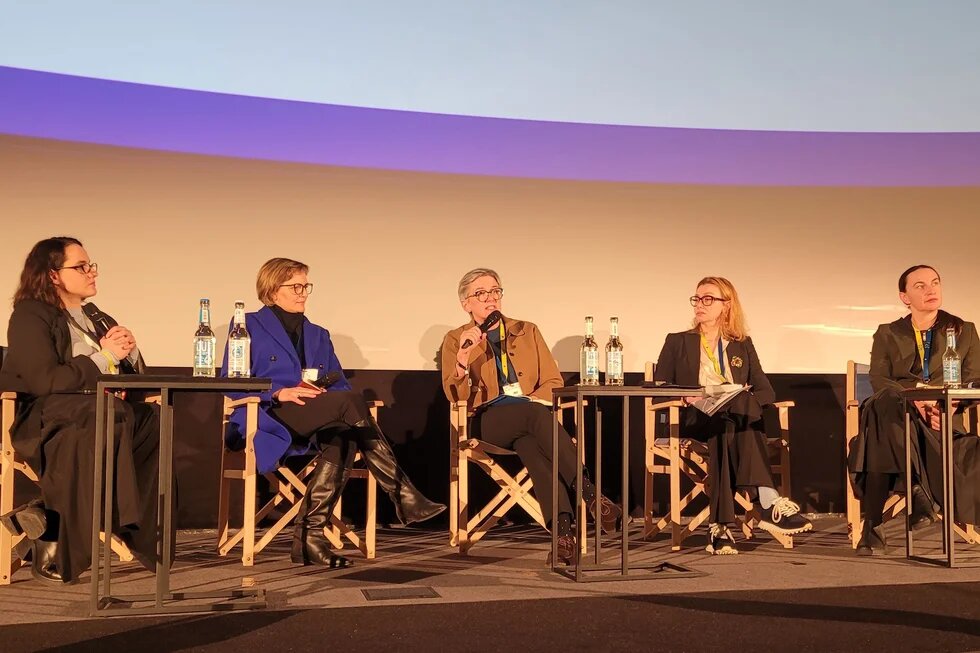 Fünf Personen sitzen auf Bühne bei Podiumsdiskussion. Mittlere Person spricht ins Mikrofon; kleine Tische mit Wasserflaschen davor.