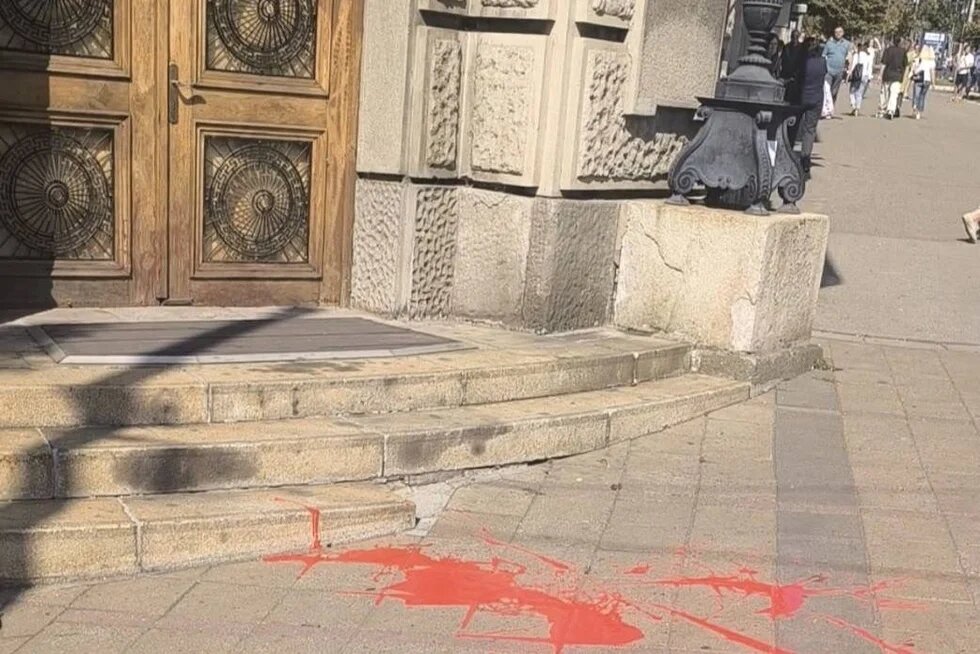 Red paint on ground in front of building steps; door and ornate facade left, pedestrians in background right.