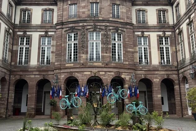 Innenhof mit historischem Gebäude; vor Bögen stehen Flaggen. In der Mitte runde Beetfläche mit mehreren aufgestellten Fahrrädern.