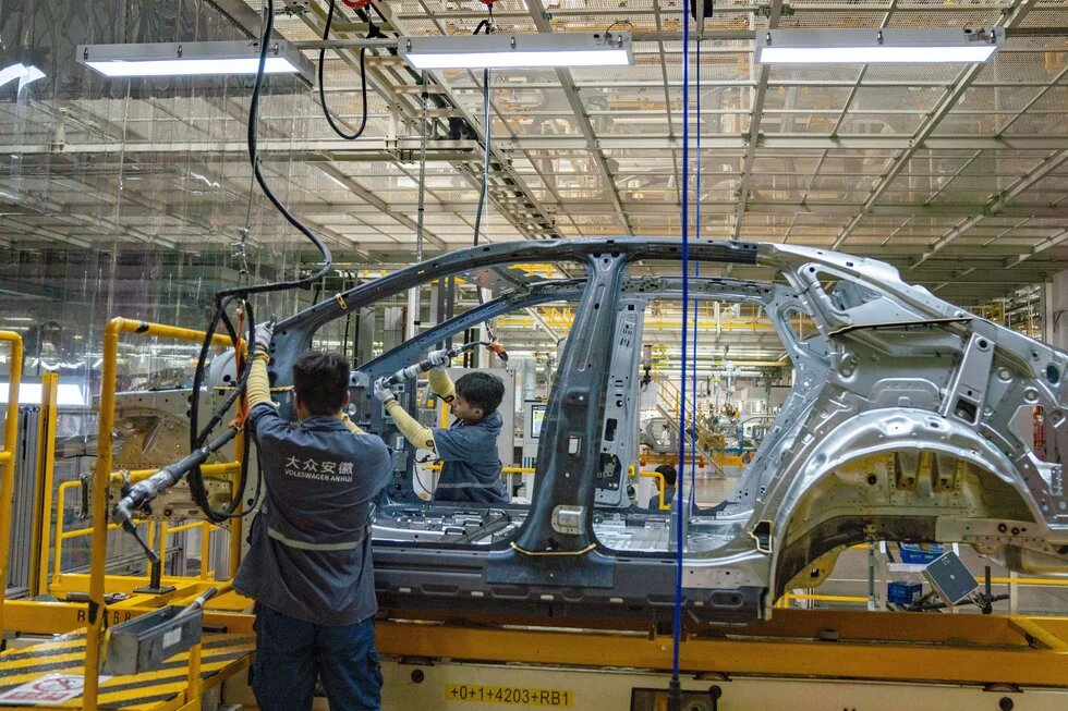 Two workers assemble a car body on a production line in a factory; cables and tools hang around, industrial hall in background.