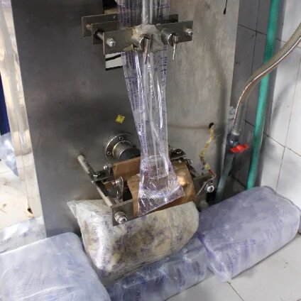 Machine fills and seals long plastic bags with liquid; several filled bags lie below, hoses and metal parts visible.