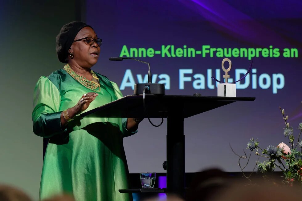 Person steht am Rednerpult und spricht; auf Leinwand dahinter Text „Anne-Klein-Frauenpreis an Awa Fall Diop“, daneben steht ein Preisobjekt.