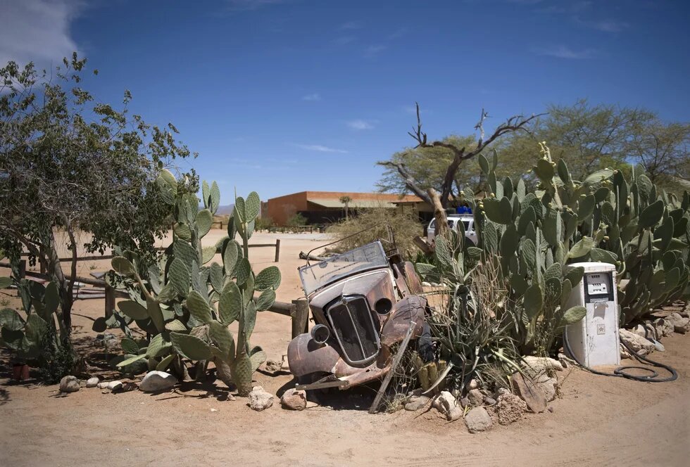 A rusty car, partially buried in the sand, stands crookedly among cacti and rocks in a dry desert landscape in front of a low building under a blue sky.