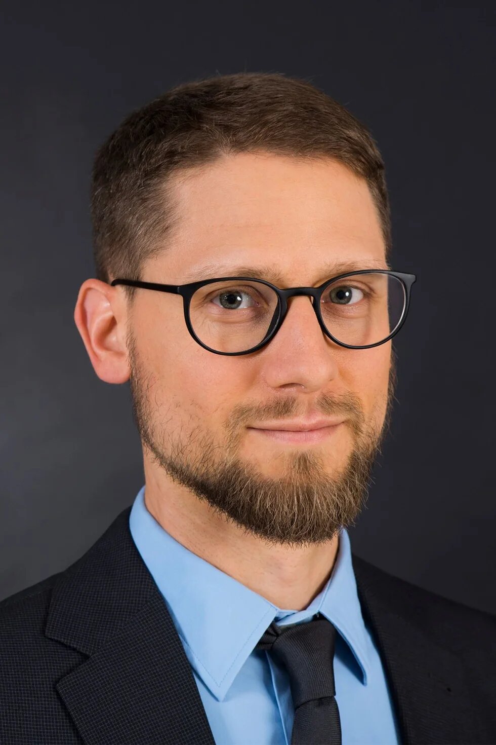 Person mit Brille und Bart blickt zur Kamera, trägt Hemd, Krawatte und Sakko, neutraler Hintergrund