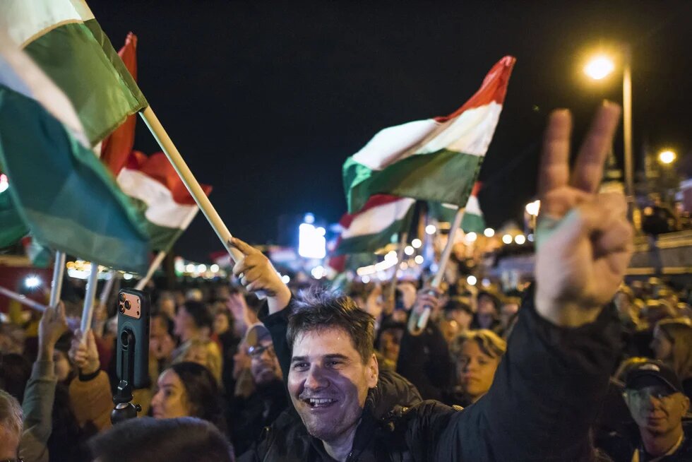 Menschenmenge nachts, mehrere halten rot-weiß-grüne Fahnen, eine Person vorne zeigt V-Zeichen, viele stehen dicht beieinander.