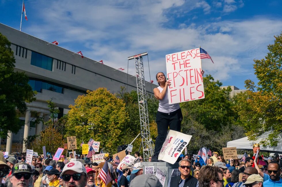 A woman stands on a platform in front of a large building, holding a sign that reads “Release the Epstein Files,” while a crowd of people around her demonstrates, carrying other signs and flags.