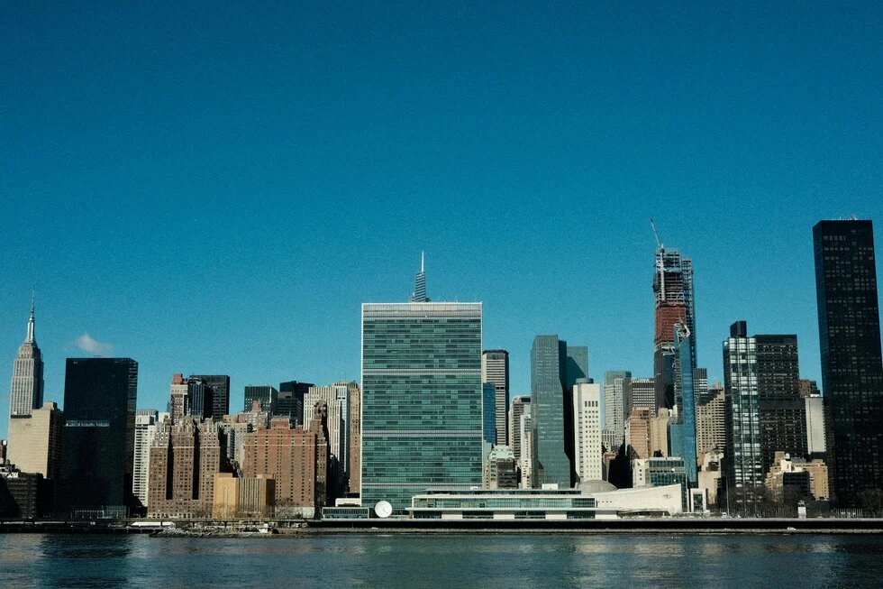 A clear city skyline with many skyscrapers, including a striking, low-rise glass building by the water in the foreground and a cloudless blue sky in the background.