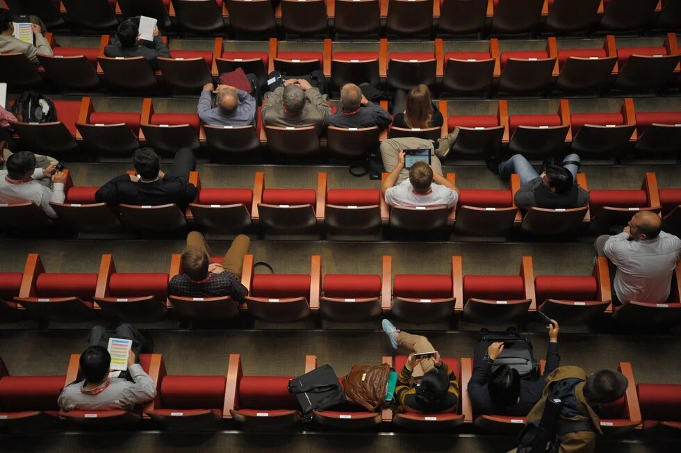 People sit scattered across rows of red seats in an auditorium, some reading or using phones and tablets.