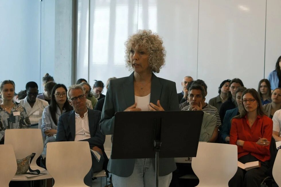 Frau mit Locken spricht an Rednerpult vor sitzendem Publikum in hellem Raum.