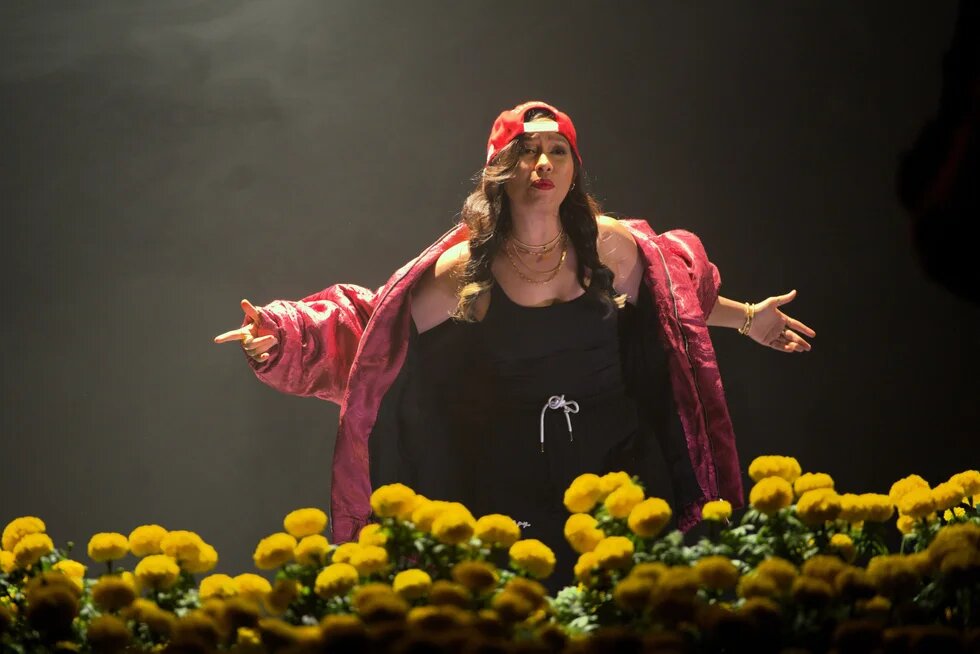Person in red cap and open jacket stands on stage behind yellow flowers, arms spread wide.