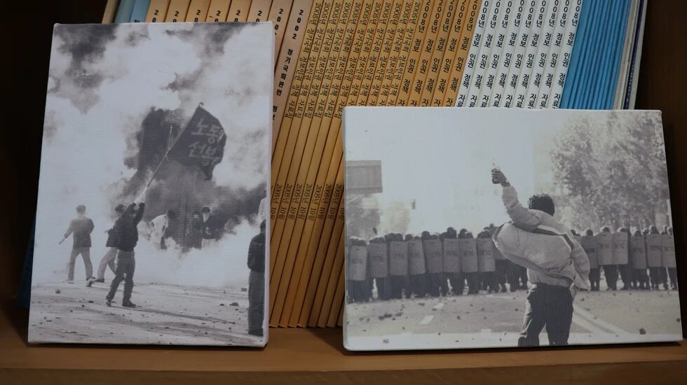 Two black and white pictures of the korean democracy movement leaning against a stack of magazines on a bookshelf.