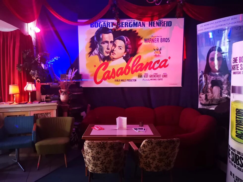 Sitzecke mit Sofa aus rotem Samt sowie Sessel im Foyer eines Kinos, an der Wand geradezu ein Plakat des Films "Casablanca", an der rechten Wand weitere Filmplakate.