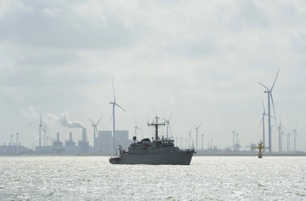 Schiff auf ruhigem Wasser, dahinter Windräder und Industrieanlagen; aus einem Schornstein steigt Rauch auf.