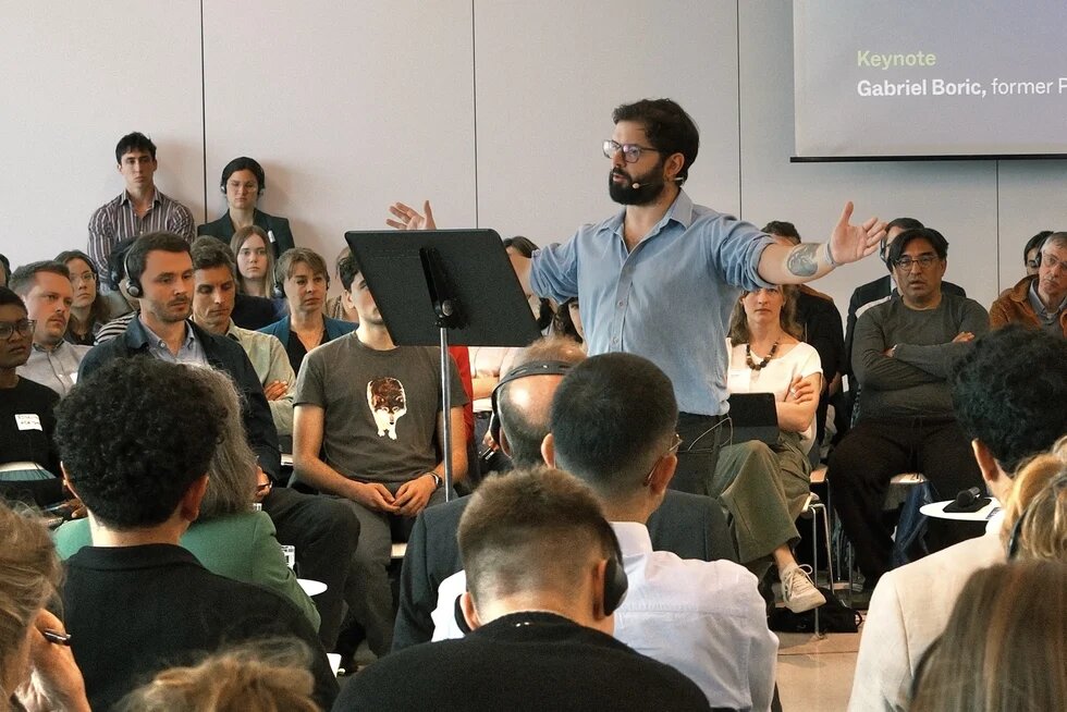 Person speaking to an audience in a conference room, screen shows “Berlin Forum on Global Cooperation”.
