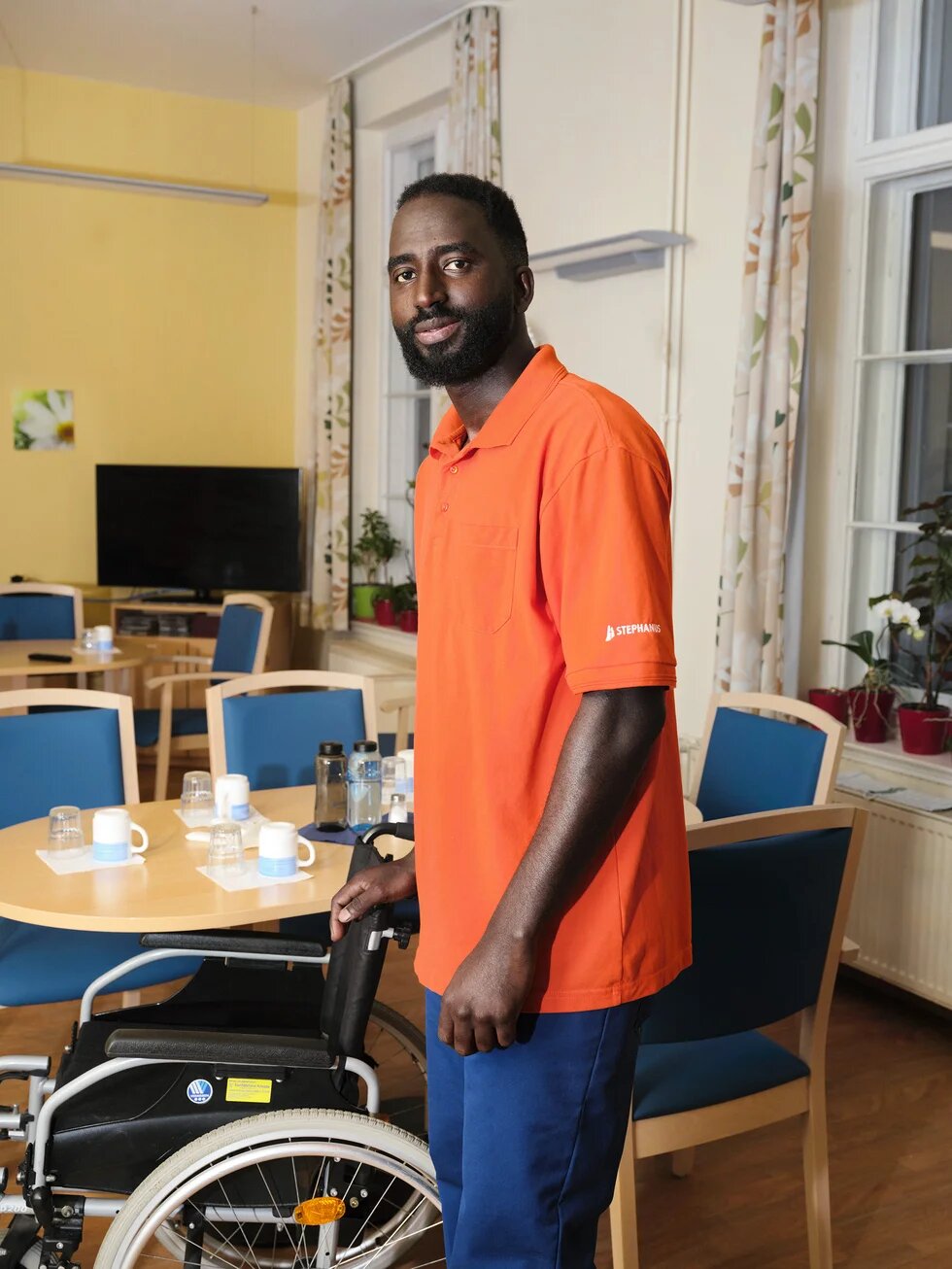 Porträt von Manjang im Essraum eines Pflegeheims. Er schiebt einen Rollstuhl.