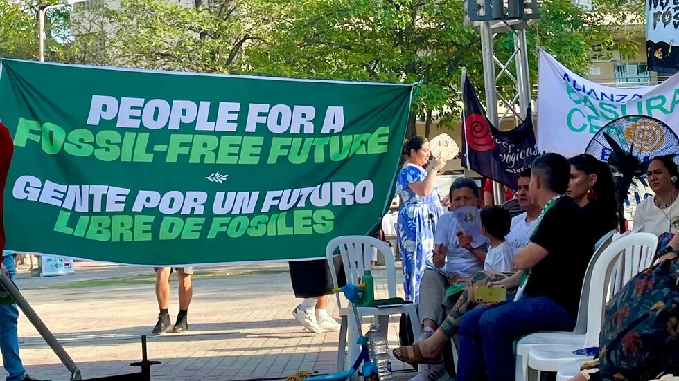 Menschen bei Kundgebung im Park. Großes Banner: „Menschen für eine Zukunft ohne fossile Energien“; mehrere Personen sitzen daneben.