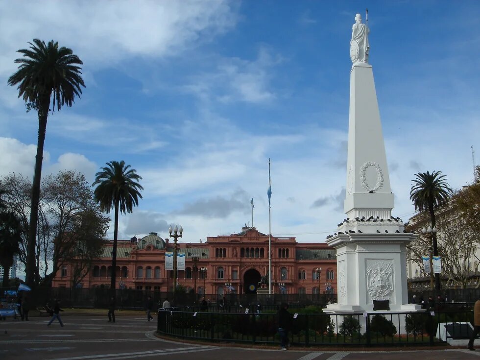 Obelisk mit Statue vor dem rosafarbenen Regierungsgebäude Casa Rosada, Palmen, Flaggen und bewölkter Himmel.