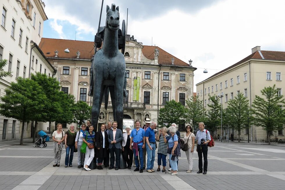Etwa 15 Personen stehen vor einer großen Reiterstatue auf einem Platz, im Hintergrund historische Gebäude und Bäume.