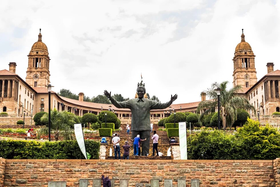 Statue von Nelson Mandela vor den Union Buildings in Pretoria, mit Gartenanlage, Menschen und zwei markanten Türmen im Hintergrund.
