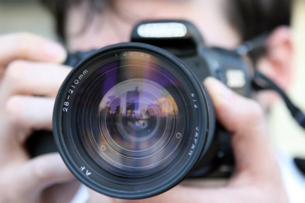 A close-up of a camera lens; the camera is being held by a person whose face is not clearly visible. The outline of a city is reflected in the lens.
