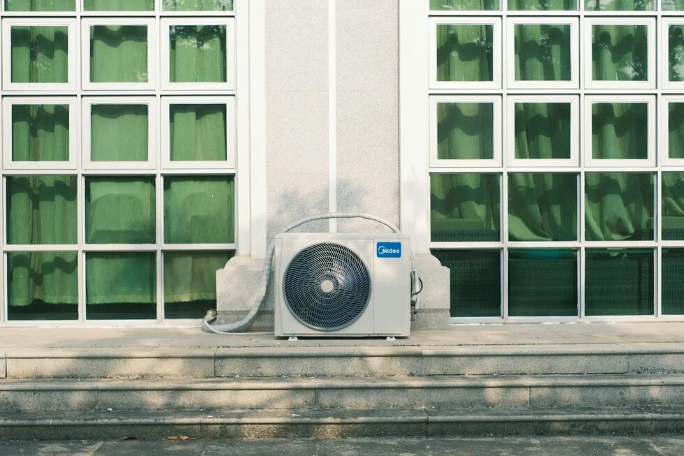 Air conditioning unit in front of a building facade between large windows with curtains; steps run along the front.