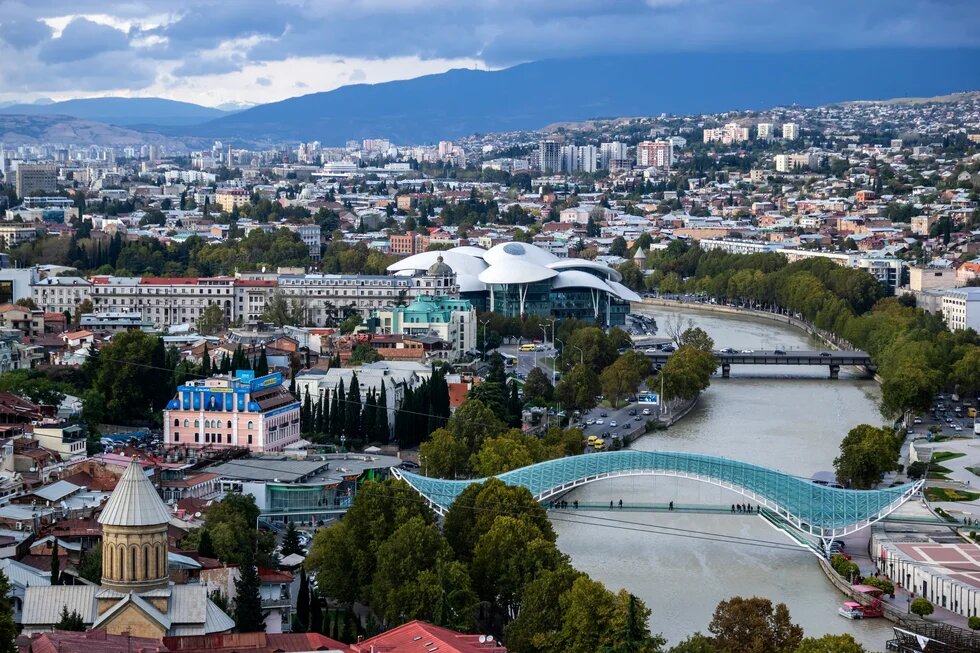 Blick über Tiflis mit Fluss, Fußgängerbrücke, markantem Kuppelbau, dicht bebauter Stadt und Bergen im Hintergrund.