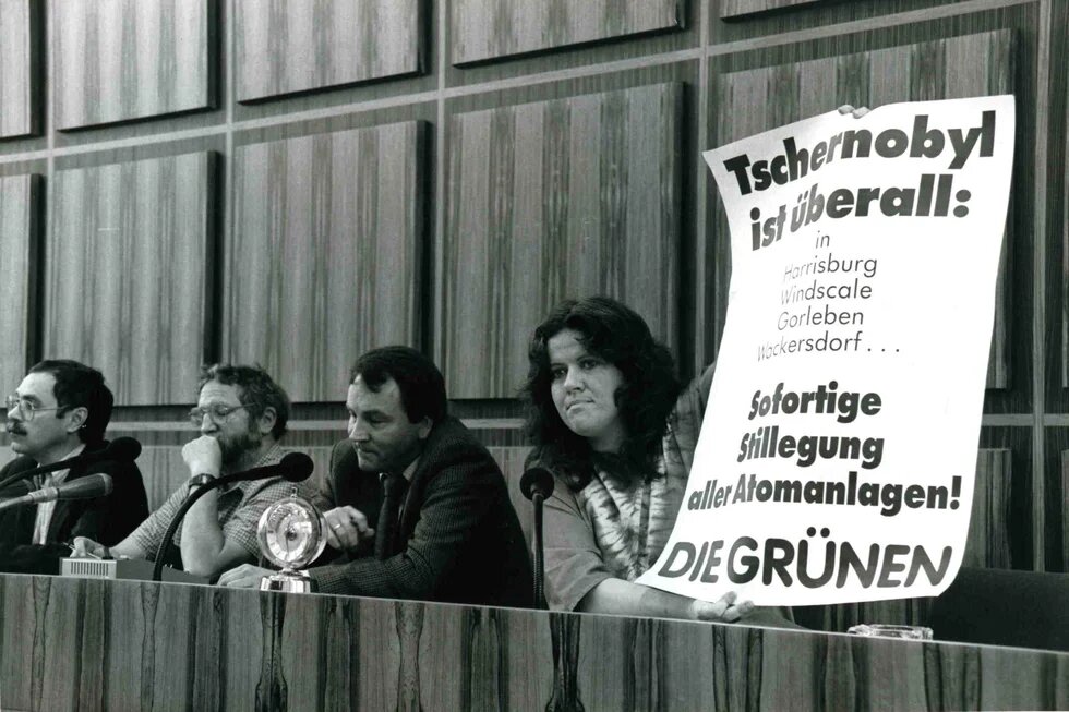 Person hält ein beschriftetes Plakat hoch, links daneben drei weitere  Personen.
