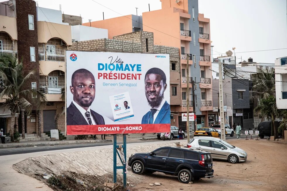 Wahlplakat mit Bassirou Diomaye Faye und Ousmane Sonko vor Wohnhäusern; davor Straße, parkende Autos und offener Entwässerungsgraben.