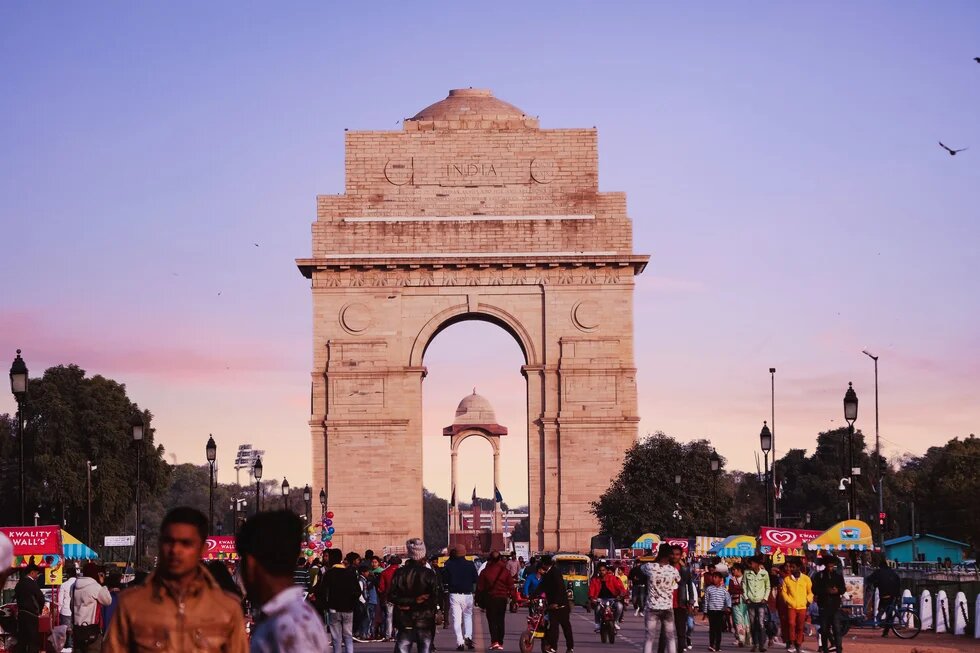 India Gate in Neu-Delhi bei Abendhimmel, davor eine belebte Straße mit vielen Menschen, Fahrzeugen und Verkaufsständen.