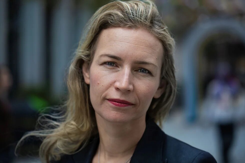 Porträt einer blonden Frau, die mit leicht geneigtem Kopf in die Kamera blickt. Im Hintergrund sind unscharf Menschen zu sehen.
