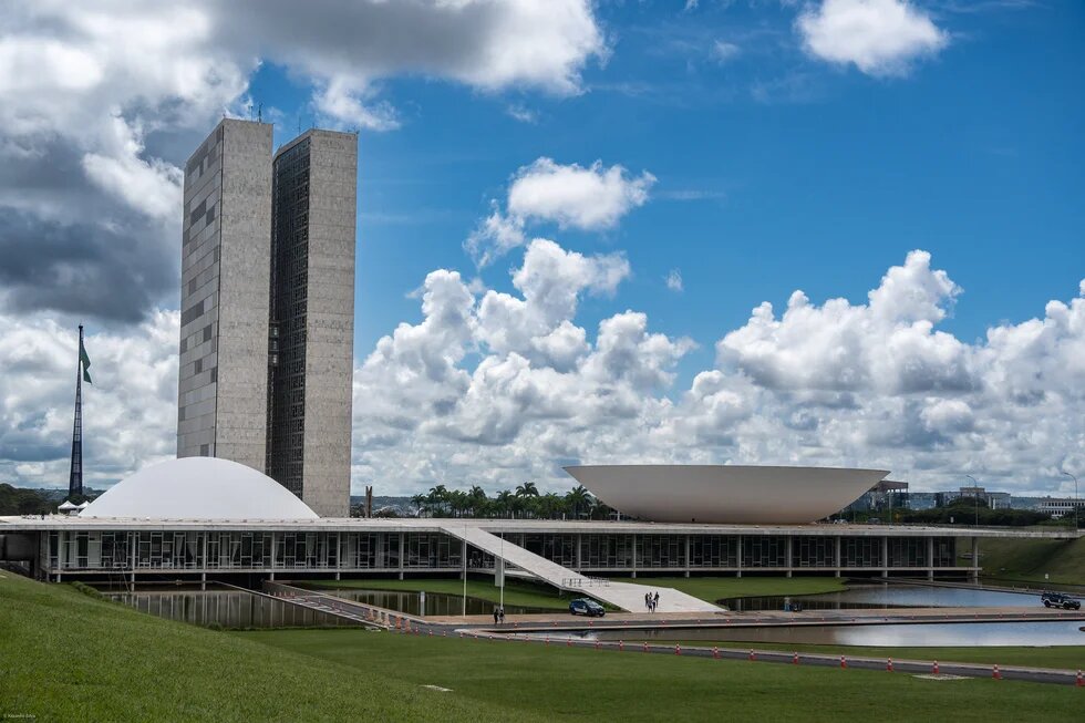 Modern government building with two tall towers and flat domed structures under a blue, cloudy sky.