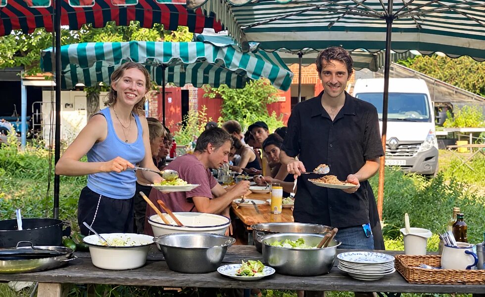 Zwei Personen stehen mit Tellern an einem Buffet im Freien; dahinter sitzen weitere Menschen unter gestreiften Sonnenschirmen.