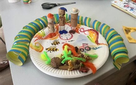 Bastelarbeit auf einem Pappteller: Figuren aus Korken, kleine Tiere und Naturmaterialien, umrahmt von einer gestreiften Papierrolle.