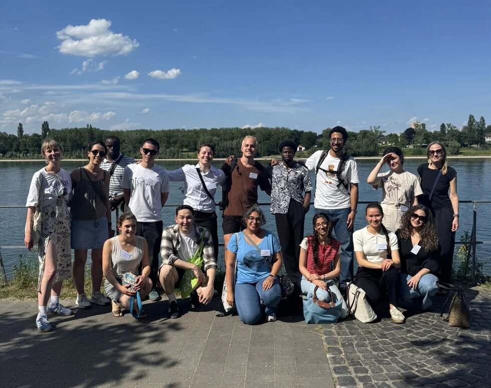 Eine Gruppe von 16 Personen posiert lächelnd am Ufer eines breiten Flusses. Im Hintergrund stehen Bäume unter blauem Himmel.