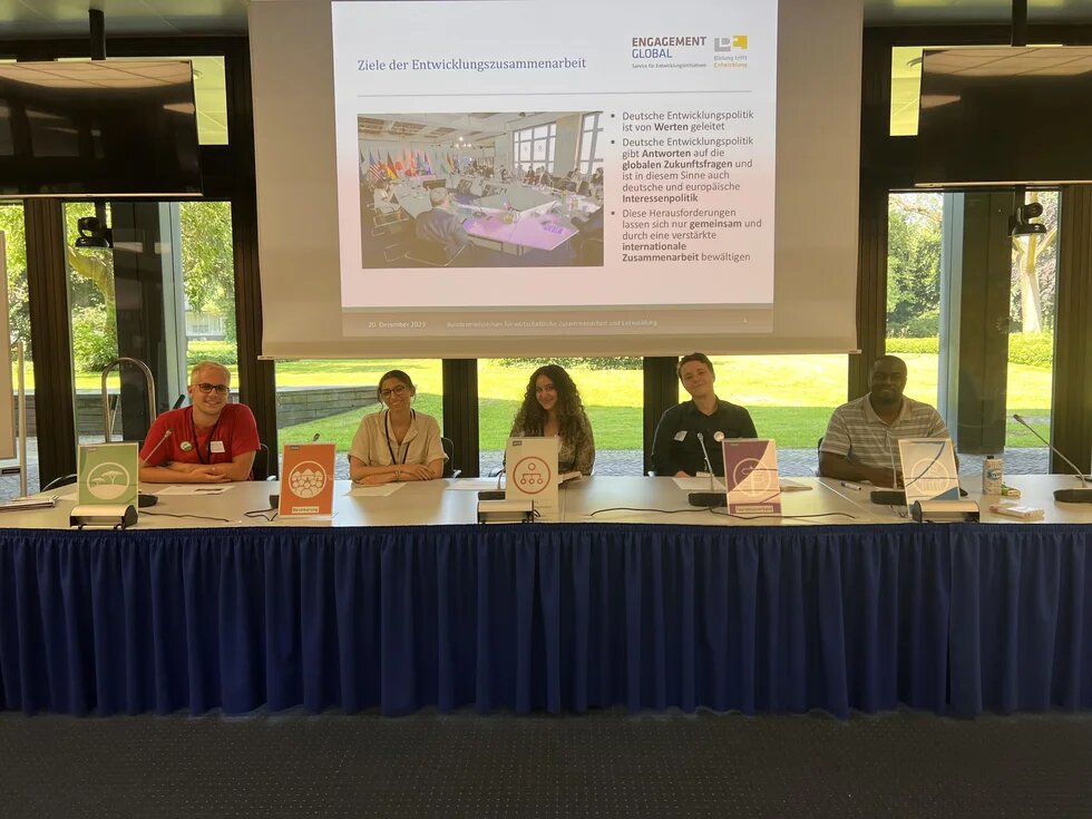 Fünf Personen sitzen an einem Konferenztisch. Dahinter zeigt eine Leinwand eine Präsentation zu Zielen der Entwicklungszusammenarbeit.