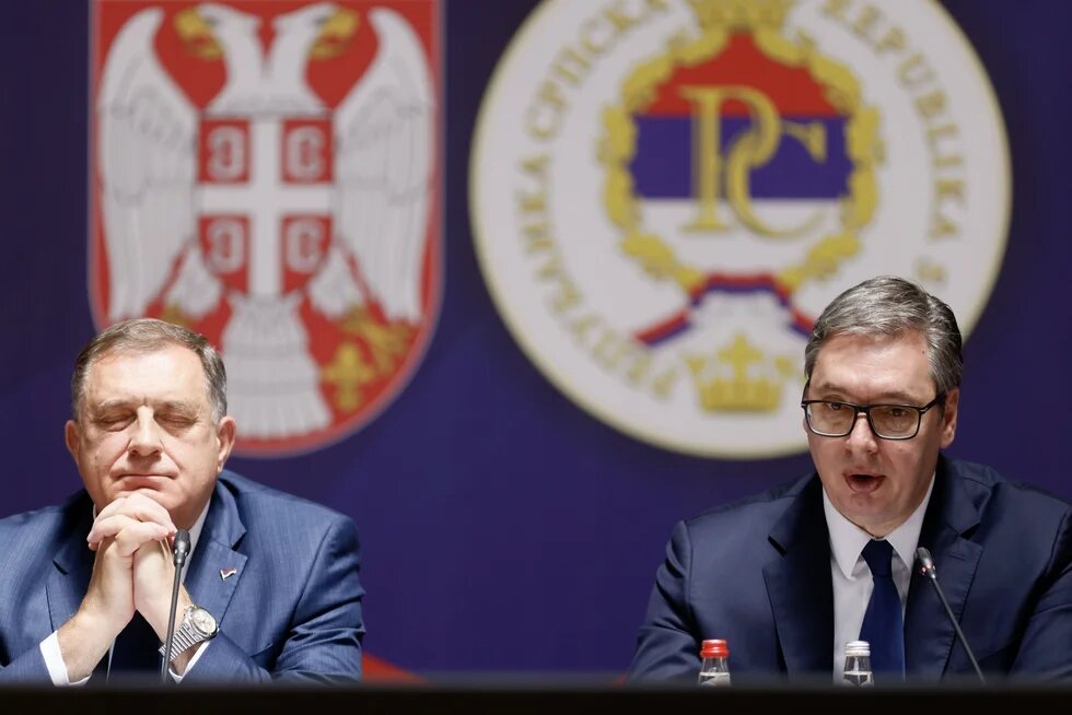 Two politicians sit at a press conference in front of Serbian state symbols. The man on the right speaks while the man on the left listens with his eyes closed.