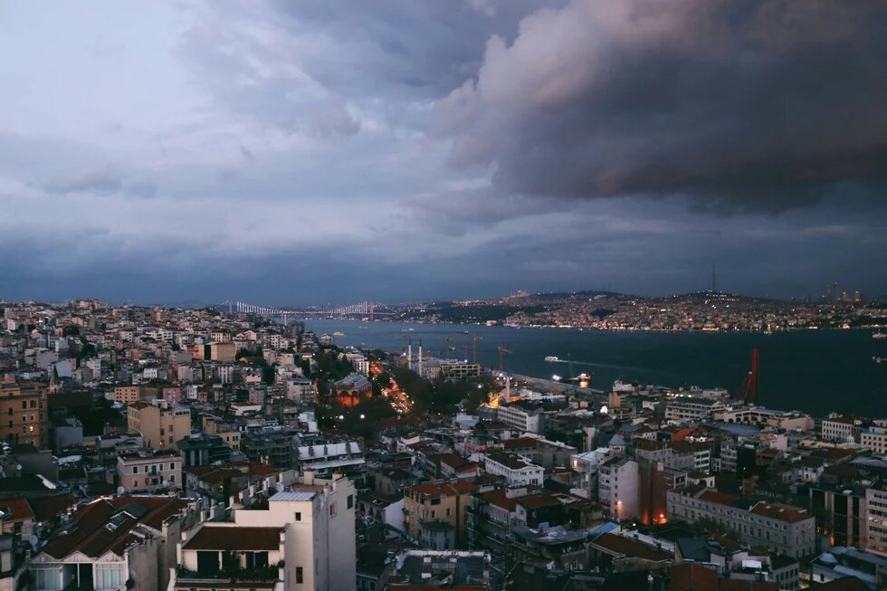Blick über dicht bebaute Stadtviertel Istanbuls auf den Bosporus und eine beleuchtete Brücke. Darüber türmen sich dunkle Gewitterwolken.