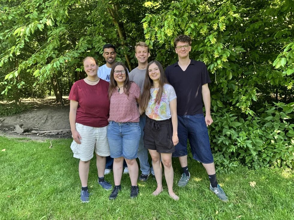 Sechs Menschen stehen für ein Gruppenfoto auf einer Wiese vor Bäumen.