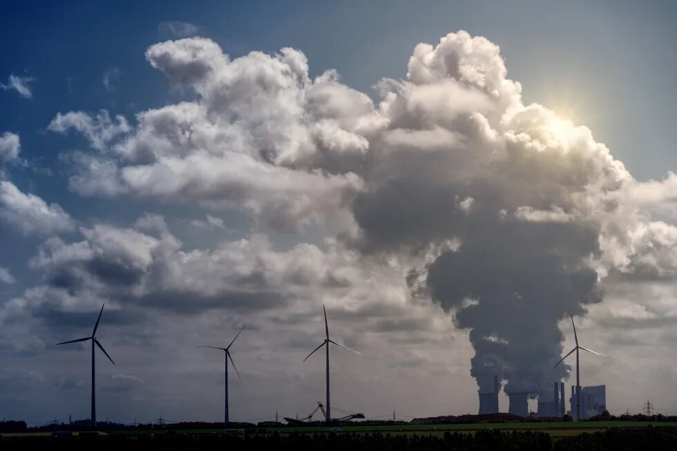 Windkraftanlagen und ein Kraftwerk unter dramatischen Cumuluswolken in einer ländlichen Landschaft.
