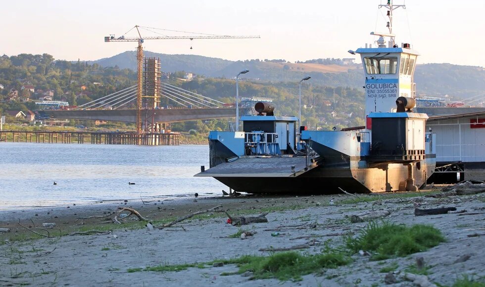 A ferry on a sandy riverbank; in the background, a bridge with a crane and stay cables is under construction.