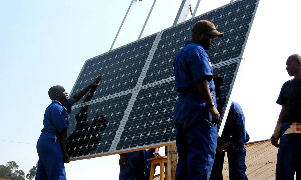 Several men in blue work overalls are setting up a large solar panel installation outdoors.