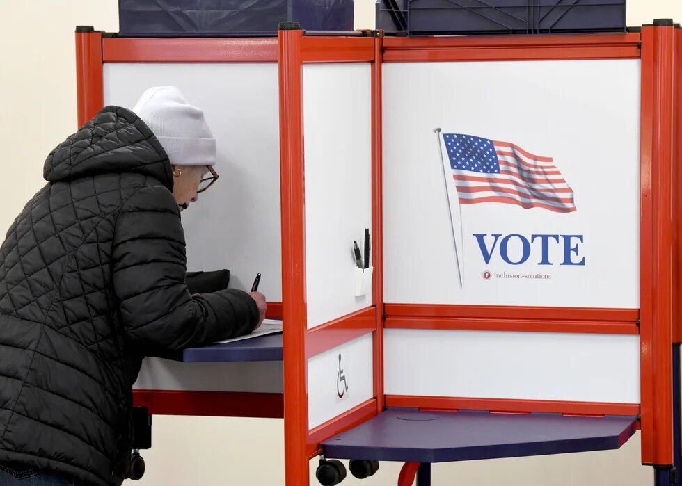 Eine Frau füllt in einer Wahlkabine einen Stimmzettel aus. An der Trennwand sind die US-Flagge und das Wort „VOTE“ (Wählen) zu sehen.