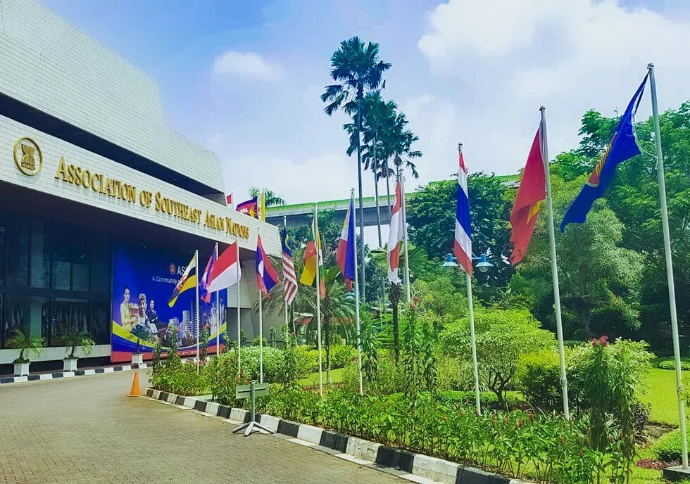ASEAN-Gebäude mit Fahnen der Mitgliedsländer vor dem Eingang, umgeben von Gartenanlage und Palmen.