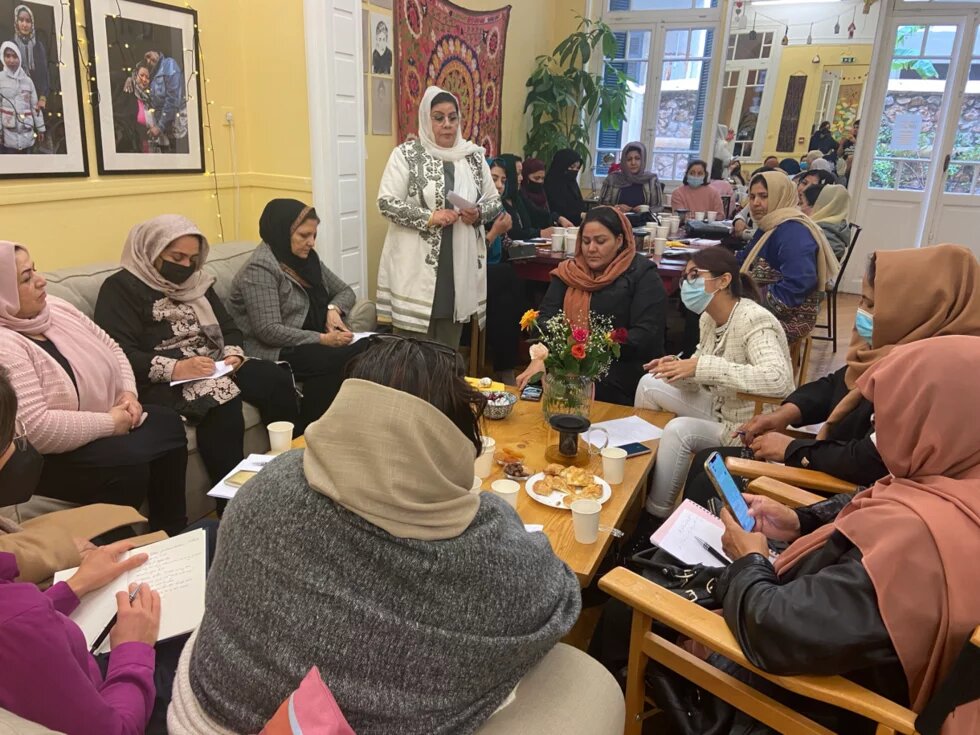 Gruppe von Frauen* in einem Raum, teils mit Notizbüchern. Eine Person steht sprechend, andere sitzen im Halbkreis. Getränke und Gebäck auf dem Tisch