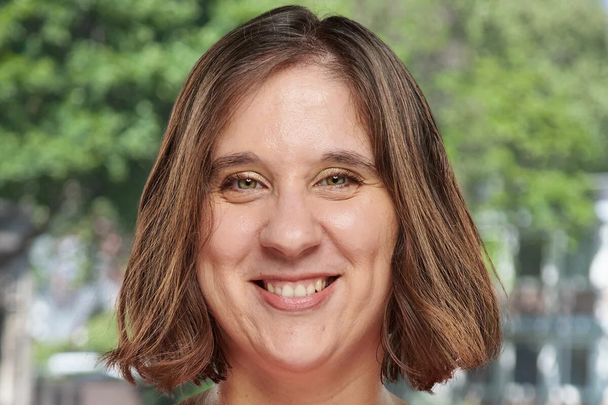 Nahaufnahme einer lächelnden Frau mit kurzen braunen Haaren. Hintergrund zeigt unscharfe, grüne Bäume.